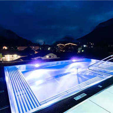 A modern infinity pool on a roof with glowing lights. In the background, mountains and a lit city can be seen.