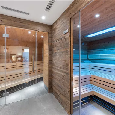 A modern wellness area with two wooden saunas. The saunas have large glass windows and soft, colored light.