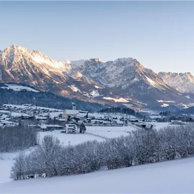 A winter landscape with snow-covered mountains and a small village in the foreground. The sun shines on the peaks, creating a bright, clear atmosphere.