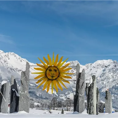 A group of stones with a sunshine-like sun in the foreground. In the background, there are snow-covered mountains and a clear blue sky.