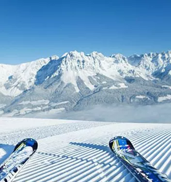 Eine schneebedeckte Berglandschaft mit klar blauem Himmel. Im Vordergrund liegen zwei Skier auf der präparierten Piste.