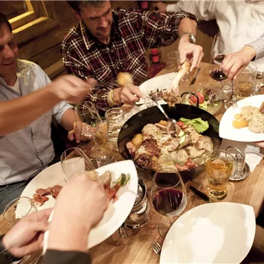 A group of people sits at a table and enjoys a lavish meal together. There are various dishes and drinks, while everyone interacts with each other.
