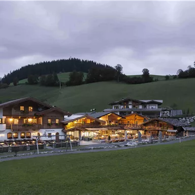 A typical alpine building at dusk, surrounded by green meadows and gentle hills. The windows emit warm light, creating an inviting atmosphere.