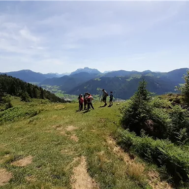 A group of people is standing on a beautiful meadow with a view of the mountains. The sky is clear and the landscape is green and hilly.