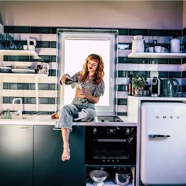 A modern kitchen with a woman sitting on the countertop pouring water from a jug. The walls are styled in elegant colors and the windows let in plenty of light.