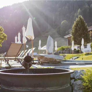 A peaceful outdoor area with lounge chairs and sun umbrellas. In the foreground is a fountain, surrounded by lush greenery and mountains in the background.