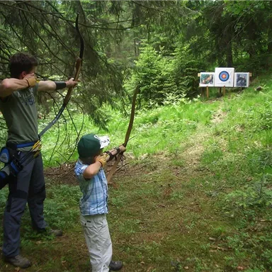 An adult and a child are practicing archery in a forest. Targets are visible in the background.