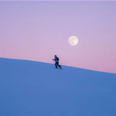 A person is hiking through a snow-covered landscape. The large, full moon shines in the sky in soft colors.