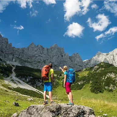 Two hikers are standing on a rock and looking at an impressive mountain landscape. The sky is clear and blue with some clouds.