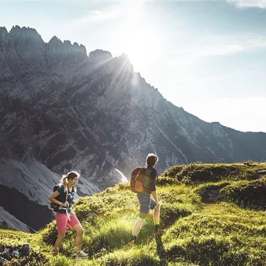 Zwei Wanderer erkunden eine grüne Wiese in den Bergen. Im Hintergrund sieht man hohe Felsen und die Sonne scheint strahlend.