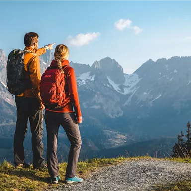 Two hikers admire the mountains in the distance. The landscape is majestic with snow-capped peaks and a clear sky.