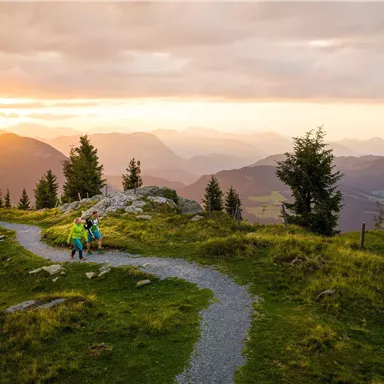 A picturesque mountain landscape at sunset with hikers on a path. The gentle hills and trees create a tranquil atmosphere.