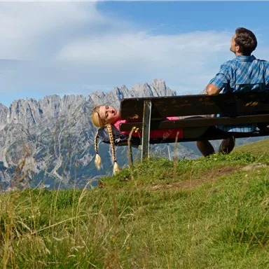 Ein Mann sitzt auf einer Bank und schaut auf die Berge. Ein Mädchen liegt entspannt auf der Bank und genießt die Aussicht.