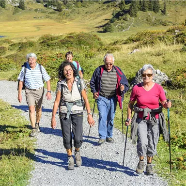 Eine Gruppe von fünf Personen wandert auf einem Schotterweg durch eine malerische Landschaft. Im Hintergrund sind grüne Hügel und ein blauer See zu sehen.