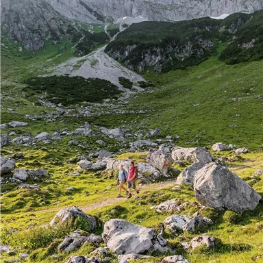 Two hikers walk through a green landscape with large stones and impressive mountains in the background. The sky is clear and nature is untouched.