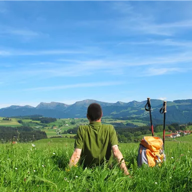 A hiker sits on a green meadow and looks at the mountains. The sky is clear and blue, in the background there are gentle hills and a small village visible.