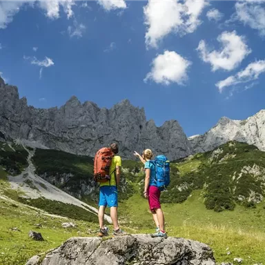 Two hikers are looking at an impressive mountain landscape. The sun is shining and the sky is clear with a few clouds.