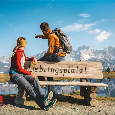 A tranquil mountain landscape with a lake in the foreground. Two hikers are sitting on a bench that reads "Favorite Spot" and enjoying the view.
