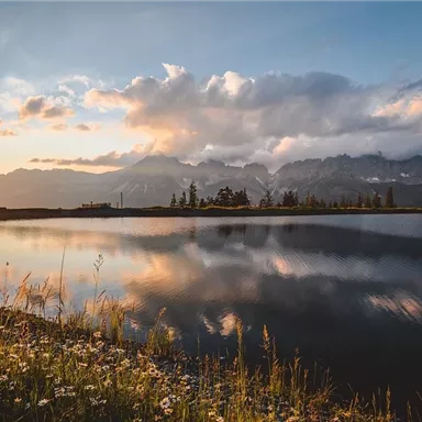 A tranquil lake with reflecting mountains in the background and colorful clouds in the sky. The gentle light of the evening sun bathes the scene in warm light.