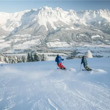 Two skiers are riding down a snowy slope in the mountains. In the background, majestic snow-capped peaks and a clear winter landscape are visible.