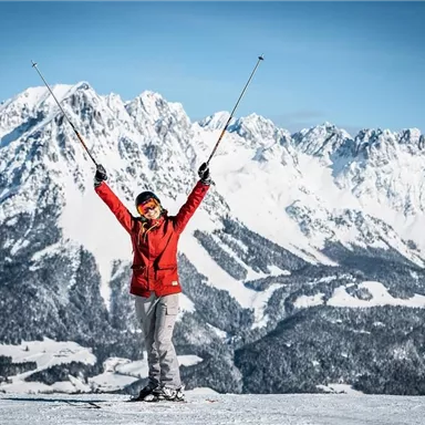A happy skier on a snowy mountain landscape raises their arms. In the background, majestic mountains and a blue sky can be seen.