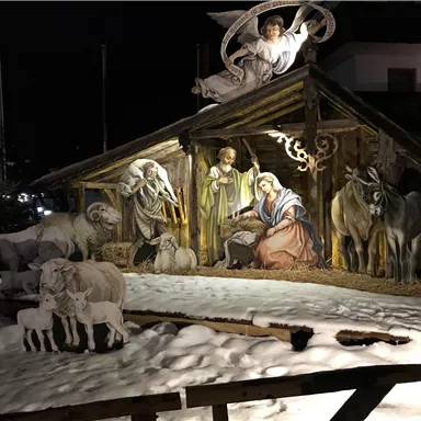 A festive nativity scene with figures of Mary, Joseph, and baby Jesus stands in a snowy environment. In the background, an angel shines, and the whole scene appears Christmas-like and contemplative.