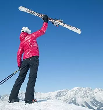 Eine Person in einem roten Skijacke hebt ein Paar Skier in die Höhe. Im Hintergrund sind schneebedeckte Berge unter einem klaren blauen Himmel zu sehen.