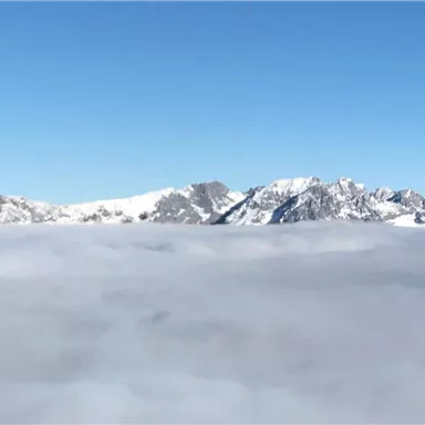 An impressive mountain landscape rises above a dense layer of clouds. The sky is clear and blue.