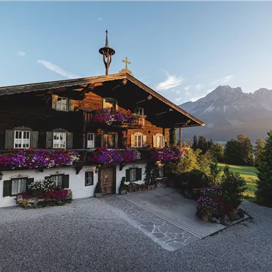 A traditional wooden house with many flower balconies. In the background, the mountains and the clear sky can be seen.