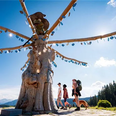 An impressive wooden sculpture in nature with radiant decorative elements. Two people and a dog are walking along a path in front of the scene.