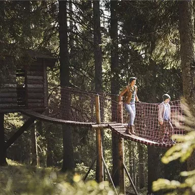 Two people are standing on a suspension bridge in the forest. In the background, a treehouse can be seen, surrounded by trees.