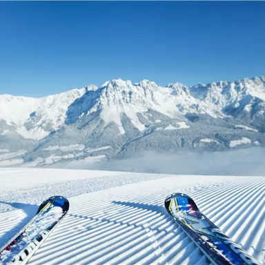 A breathtaking snow landscape with mountains in the background. Skis are lying on the freshly groomed slope under a clear blue sky.