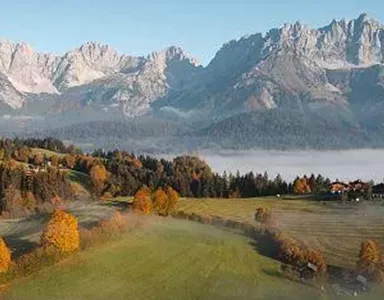 A picturesque landscape with impressive mountains in the background. In the foreground, there are green meadows and autumnal trees.