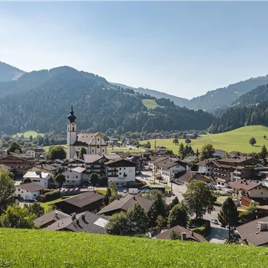 A picturesque mountain landscape with a small village and a church in the foreground. The surroundings are green with gentle hills and mountains in the background.