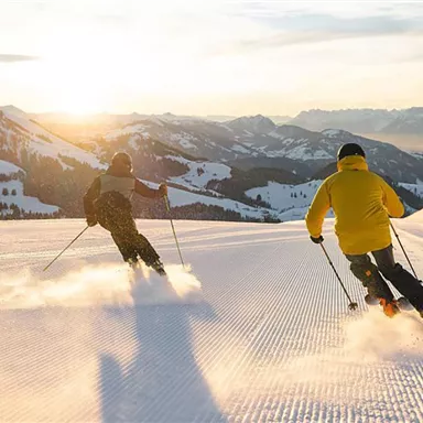 Zwei Skifahrer fahren bei Sonnenuntergang eine verschneite Piste hinunter. Im Hintergrund sind Berge und ein klarer Himmel zu sehen.