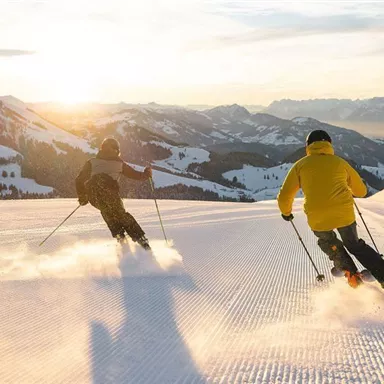 Two skiers are gliding down a snowy slope at sunset. In the background, snow-covered mountains can be seen.