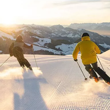 Two skiers are riding in the evening sun over a snow-covered slope. The mountains in the background provide a picturesque backdrop.