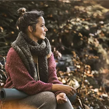 A woman is sitting in an autumn forest, observing her surroundings. She is wearing a warm sweater and a scarf.