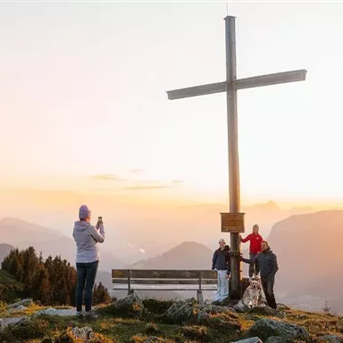 A group of people gathers around a large cross on a mountain. In the background, a picturesque mountain landscape can be seen at sunset.