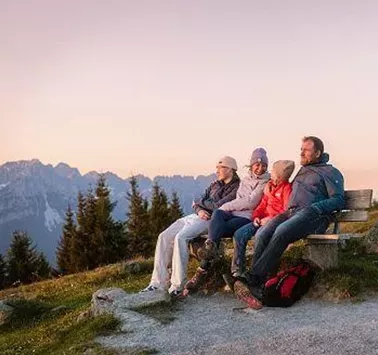 A family is sitting on a bench in the mountains, enjoying the view. The landscape is surrounded by high mountains and trees.