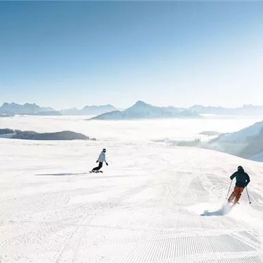 Eine sonnige Winterszene mit drei Skifahrern auf frischem Schnee. Im Hintergrund sind majestätische Berge und ein klarer Himmel zu sehen.