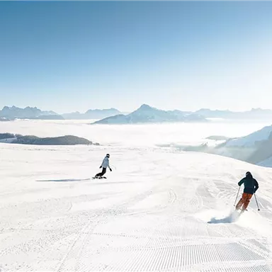 Drei Skifahrer fahren auf frischem Schnee in den Bergen. Ein klarer Himmel und weite Ausblicke schaffen eine schöne Winterlandschaft.