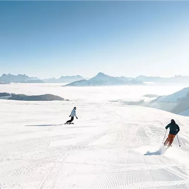 Three skiers are skiing down a snow-covered slope. The mountain landscape is clear and sunny, with a blue sky in the background.