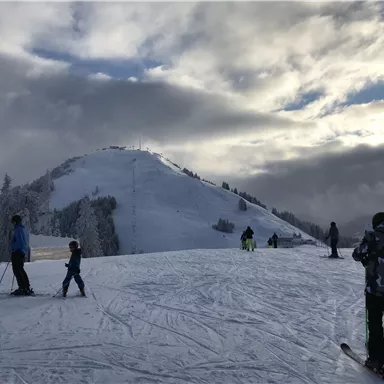 A snowy mountain landscape with skiers on the slopes. The sky is cloudy, but the light breaks through the clouds.