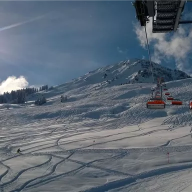 A snow-covered slope with ski tracks and a sunny, clear sky. In the background, a ski lift can be seen.