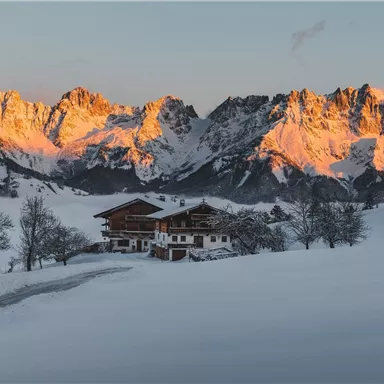 A picturesque winter landscape with snow-covered mountains in the background. In the foreground stands a cozy wooden house, surrounded by trees.