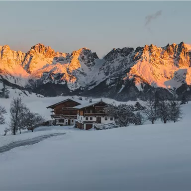 Eine malerische Winterlandschaft mit schneebedeckten Wiesen und majestätischen Bergen. Ein traditionelles Chalet steht vor den beeindruckenden, von der Abendsonne beleuchteten Gipfeln.