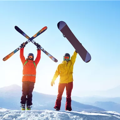 Two skiers are standing on a snow-covered summit. One is raising skis, the other a snowboard into the air.