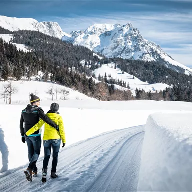 A couple is walking on a snowy path in the mountains. The surroundings are idyllic with snow-covered trees and impressive mountains in the background.