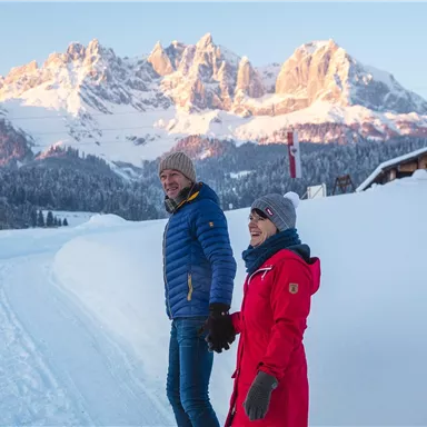 A couple walks hand in hand on a snowy path. In the background, impressive mountains and a clear sky can be seen.